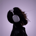 Side profile of a person wearing large over-ear AirPods Max, head tilted back, with short hair slightly flowing. The background is a soft purple gradient. The person is dressed in a dark jacket, creating a stylish, modern vibe.