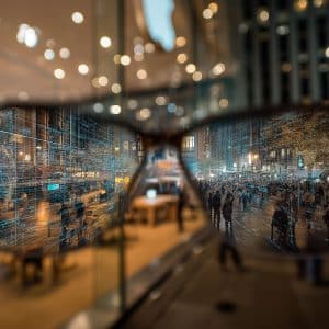 A person holds up smart glasses, through which a city street scene appears enhanced with digital data overlays. The Apple logo is visible in the lower right corner of the image.